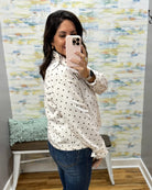 A woman taking a mirror selfie wearing a white long-sleeve blouse with black polka dots and blue jeans, standing in front of a patterned wallpapered wall.