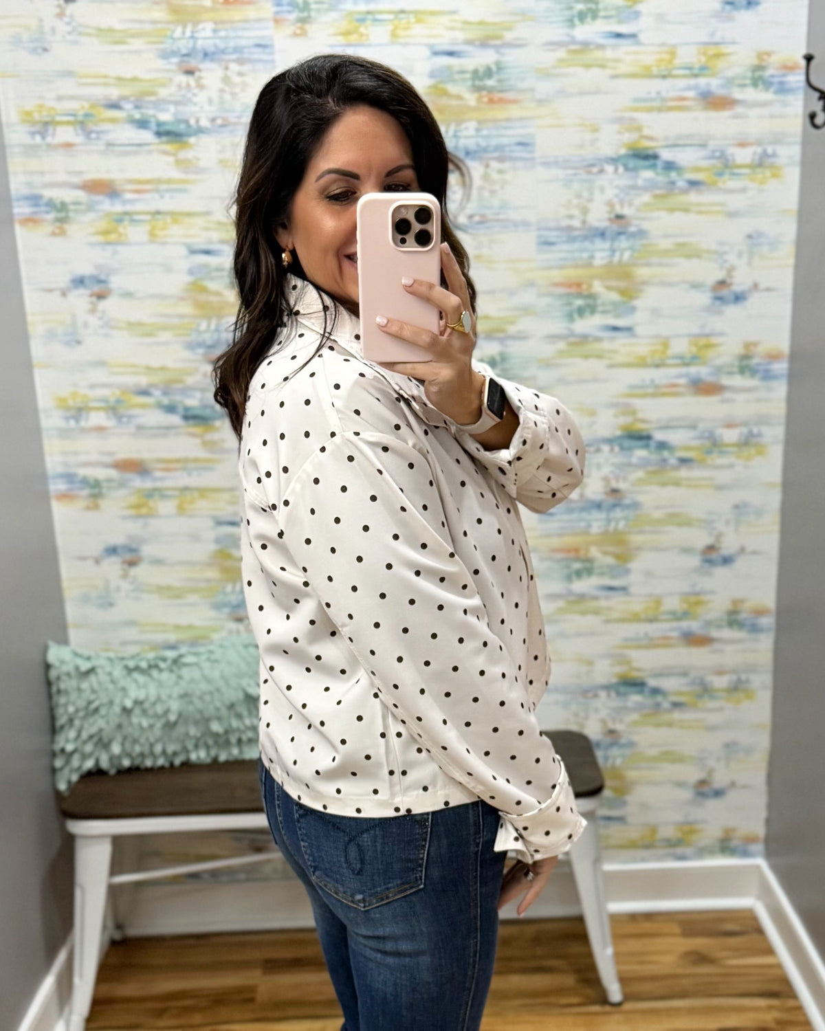 A woman taking a mirror selfie wearing a white long-sleeve blouse with black polka dots and blue jeans, standing in front of a patterned wallpapered wall.