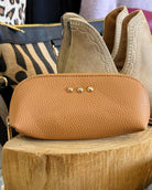 A tan pebbled leather zippered pouch with three gold-tone studs, displayed on a wooden block with other handbags in the background.