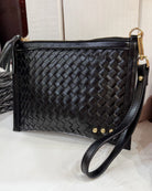 Black woven leather clutch handbag with a wristlet strap, gold-tone hardware, and a tassel detail, displayed on a light surface.