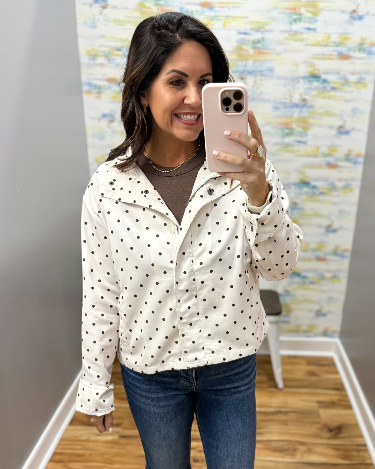A woman takes a mirror selfie wearing a cream-colored zip-up jacket with black polka dots over a brown top and blue jeans.