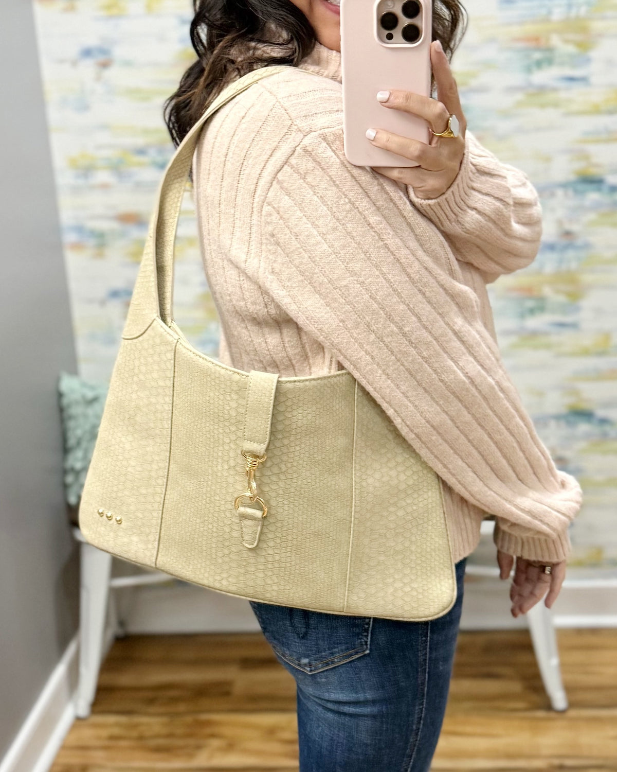 A woman takes a mirror selfie showing a light beige textured shoulder bag slouching on her shoulder. She is wearing a cream ribbed knit sweater and blue jeans, with a patterned wall and wooden floor in the background.