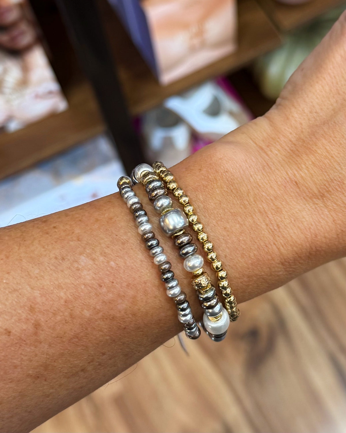 Three beaded stretch bracelets on a person's wrist, featuring silver and gold metallic beads, white faux pearls, and textured round beads.