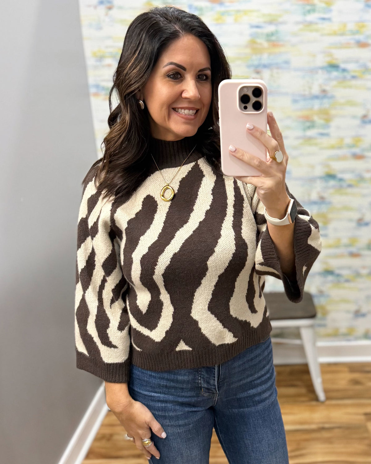 A woman taking a mirror selfie in a boutique, wearing a brown and cream zebra-patterned knit sweater and blue jeans.