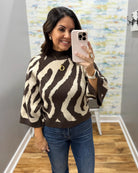 A woman takes a mirror selfie wearing a brown and cream zebra-patterned knit sweater, blue jeans, and a gold necklace. She is standing in an indoor hallway with a wood floor and a gray wall.