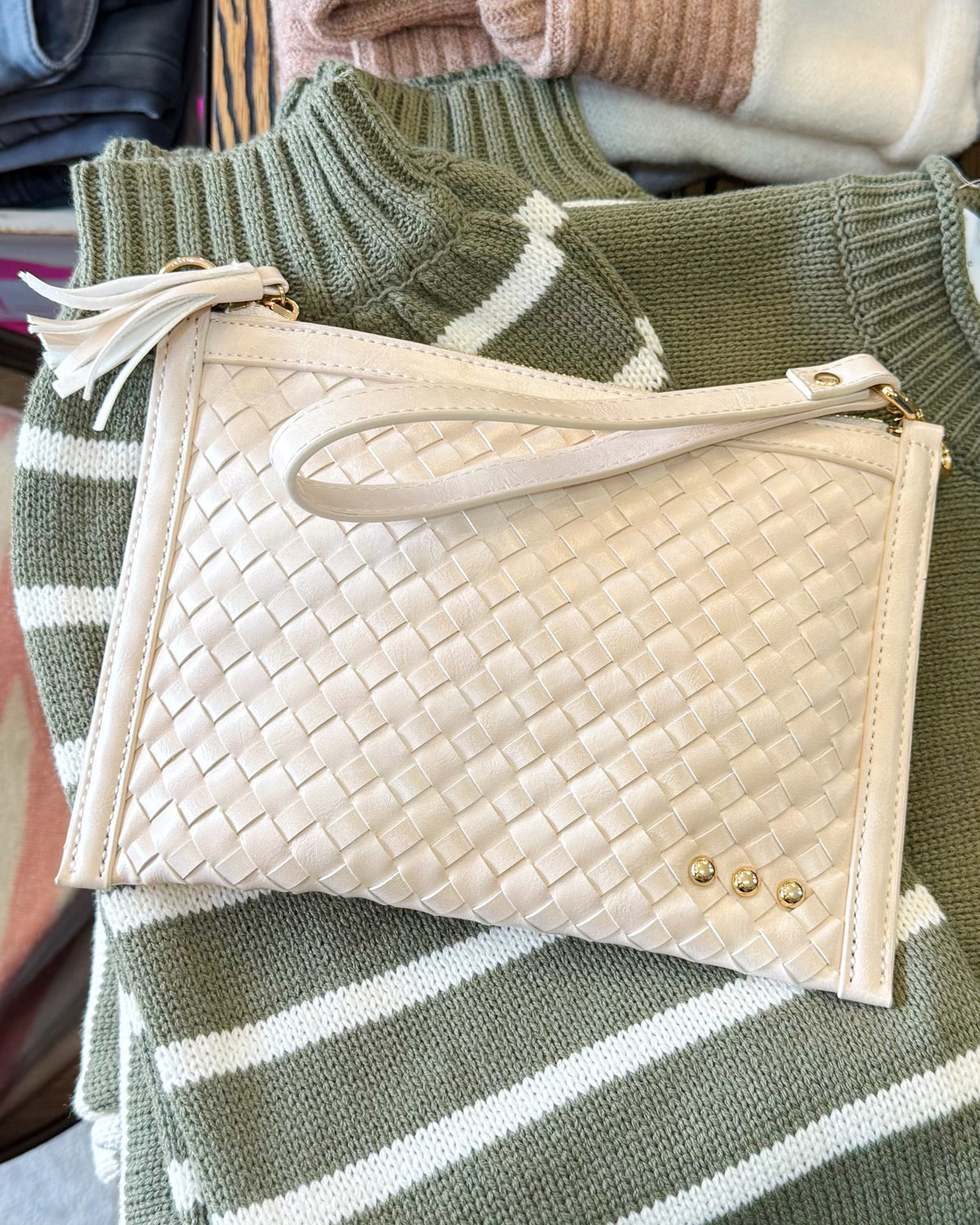 A woven cream clutch bag with a wrist strap and gold-tone studs, displayed on top of a green and white striped knit sweater.