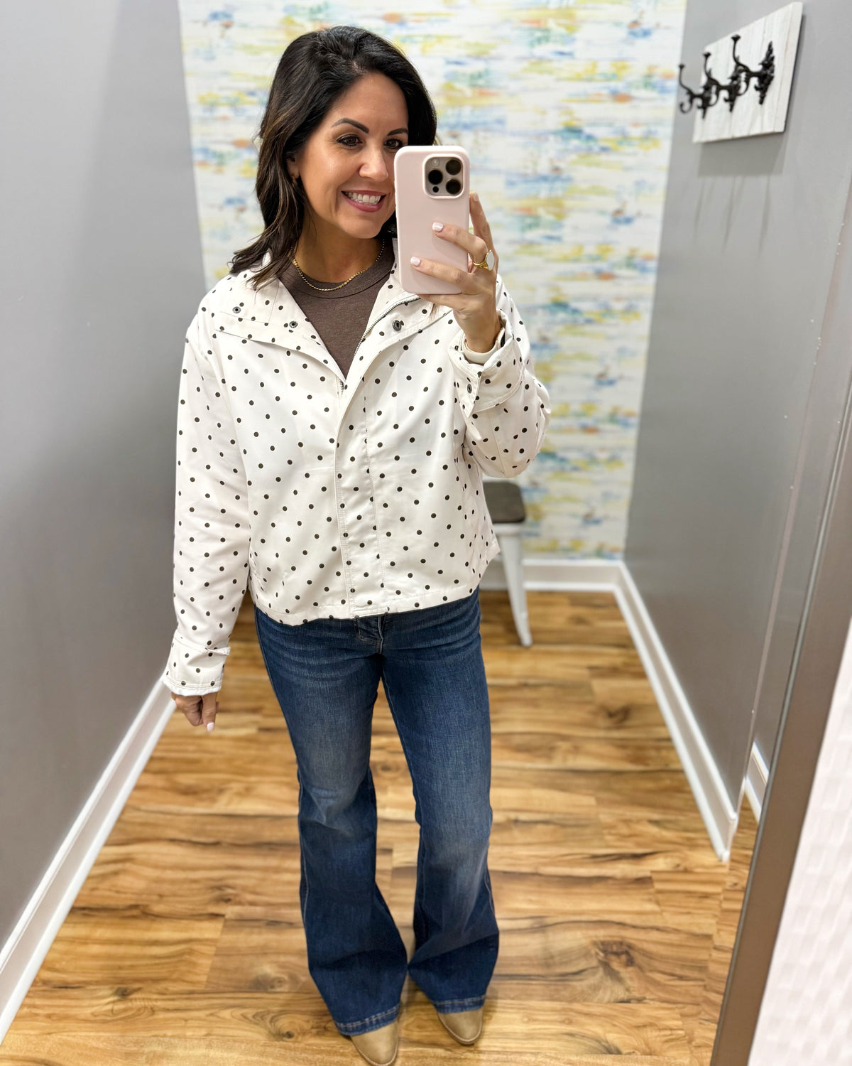 A woman takes a mirror selfie wearing a white long-sleeve button-up shirt with black polka dots, a brown crewneck undershirt, blue flare jeans, and metallic gold shoes. She is standing in a fitting room with wood floors and gray walls.