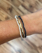 Three delicate bracelets worn on a person's wrist: a white pearl strand, a gold-toned abstract bar bracelet, and a dark navy beaded bracelet, set against a blurred wooden background.
