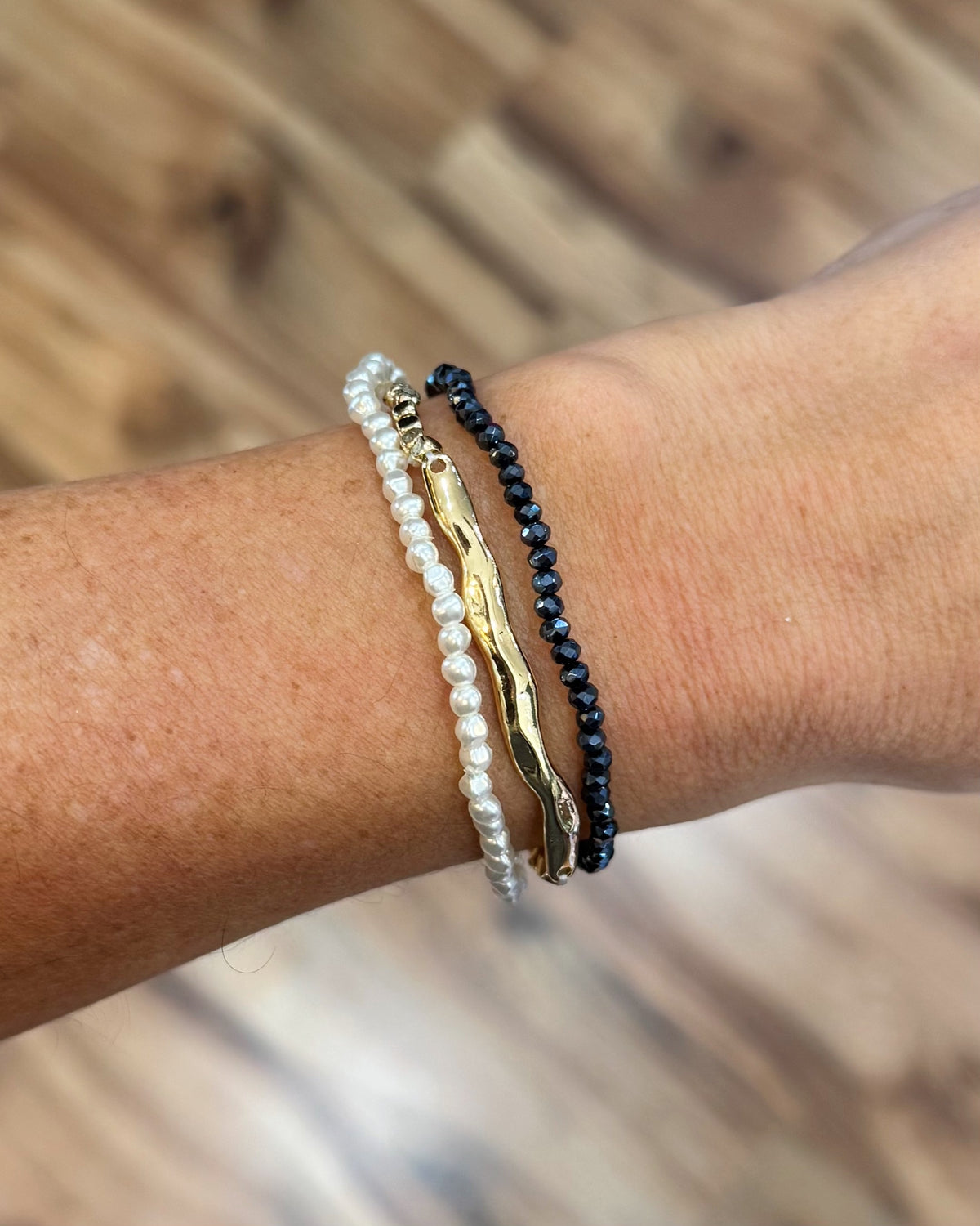 Three delicate bracelets worn on a person's wrist: a white pearl strand, a gold-toned abstract bar bracelet, and a dark navy beaded bracelet, set against a blurred wooden background.