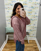 A woman takes a mirror selfie wearing a dusty rose pink ribbed knit hooded sweater and blue jeans in a fitting room with a patterned wallpaper background.