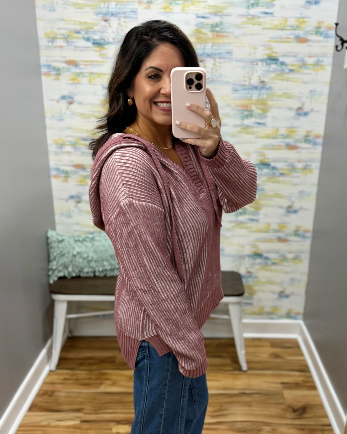 A woman takes a mirror selfie wearing a dusty rose pink ribbed knit hooded sweater and blue jeans in a fitting room with a patterned wallpaper background.
