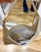 A close-up of a beige shoulder bag with a textured exterior and a patterned interior, shown hanging in front of a mirror that reflects a person's legs.