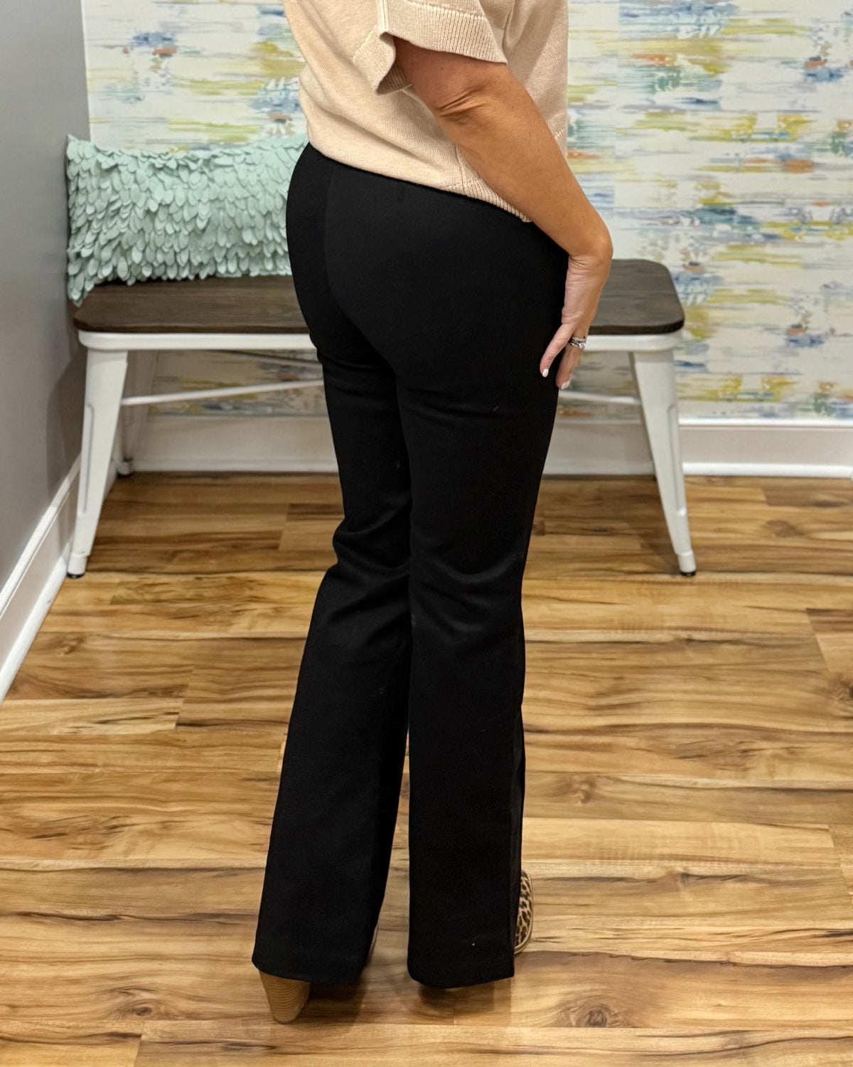 A person shown from the back wearing black flared pants and a light beige top, standing on a wood floor in front of a bench and patterned wall.