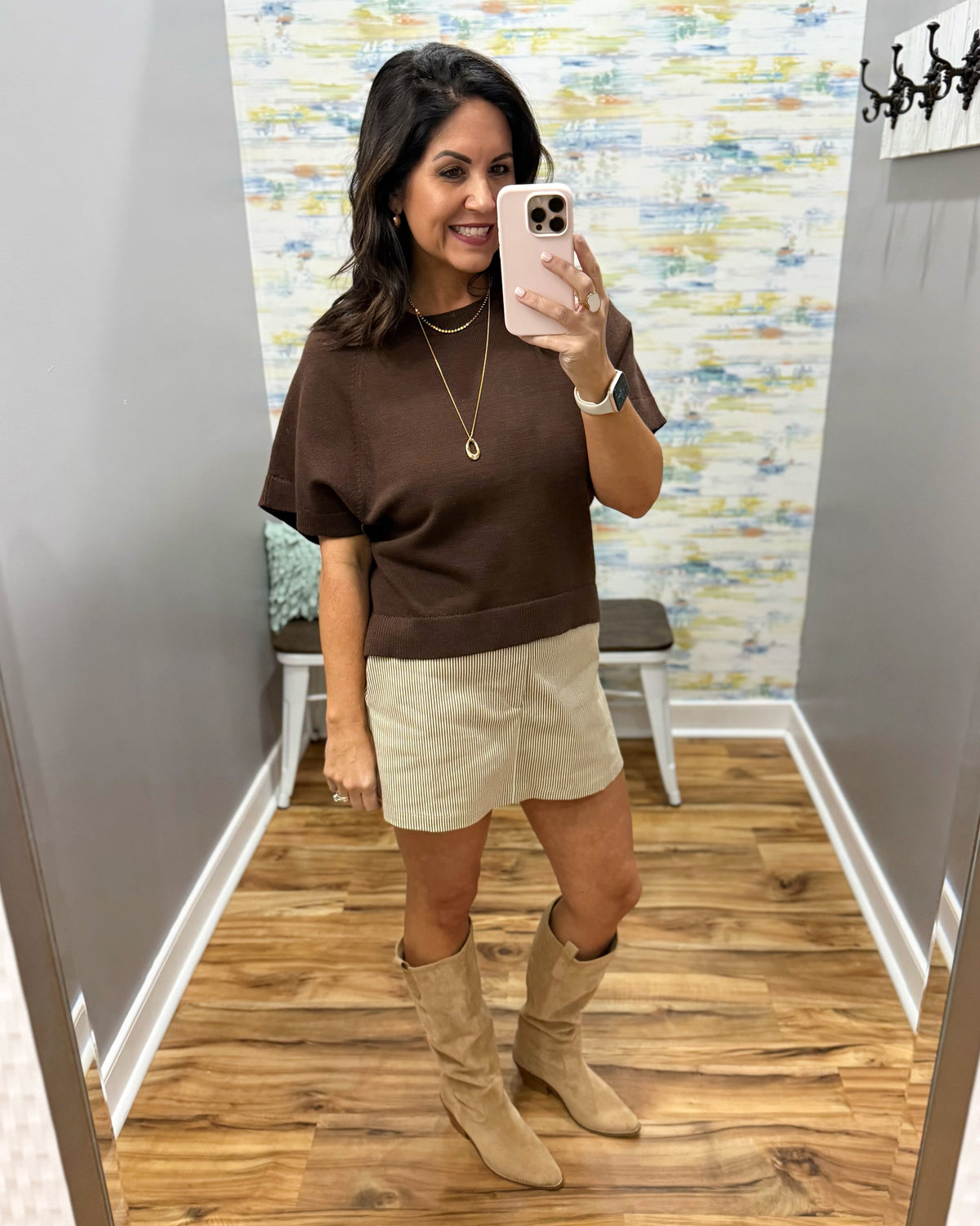 A woman takes a mirror selfie in a fitting room wearing a brown short-sleeve knit top, a beige mini skirt, and beige knee-high boots.