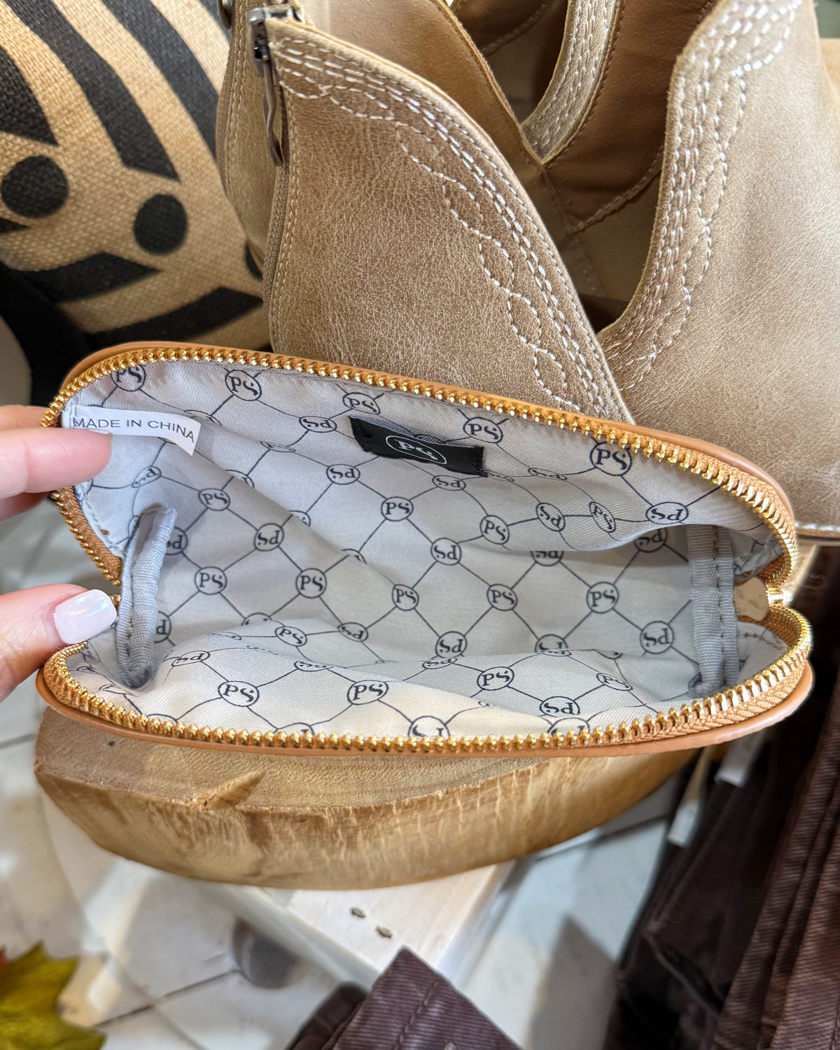 A close-up of a beige suede zippered pouch or cosmetic bag being held open by a hand, showing a patterned interior lining with a gold-tone zipper.