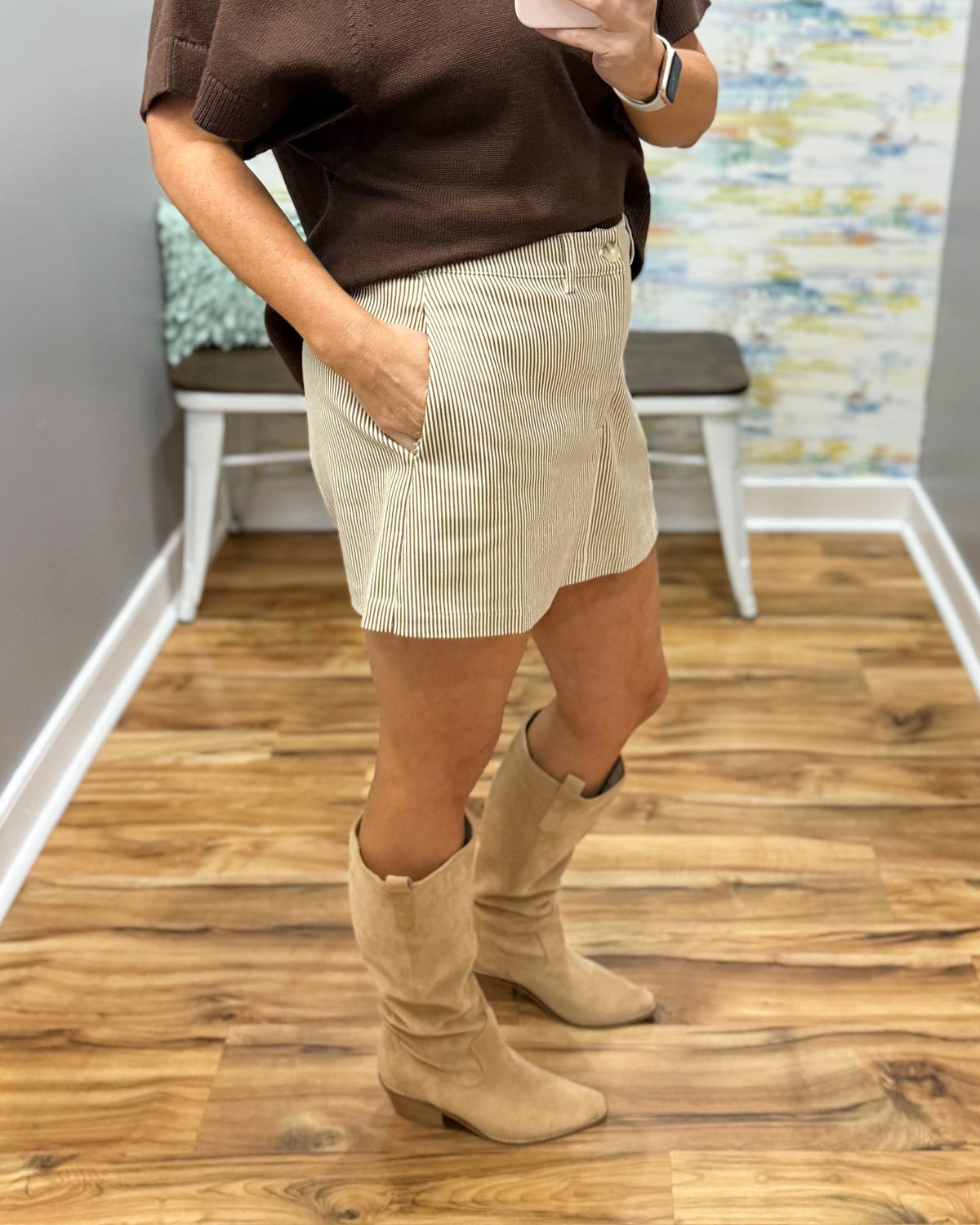 A person wearing a brown short-sleeve top, beige corduroy shorts, and beige knee-high boots, standing in a room with wood floors and a bench in the background.