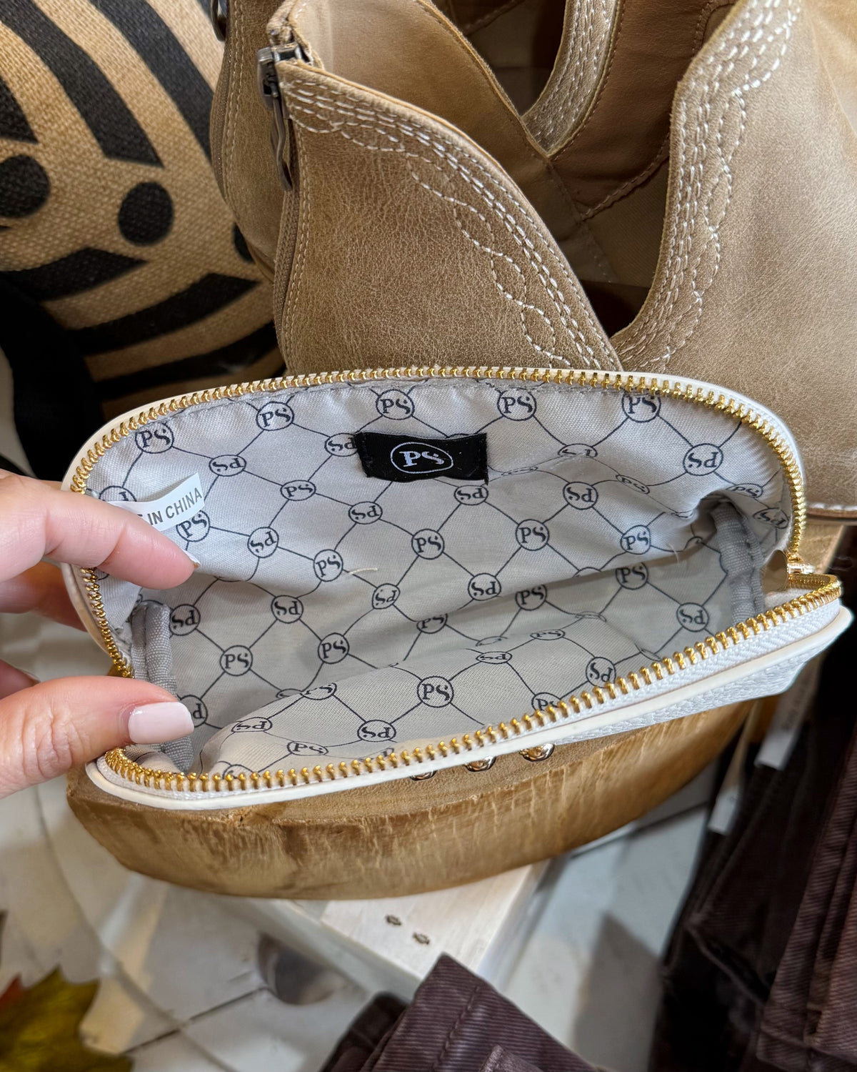 A close-up of a beige suede or faux-suede zip pouch being held open by a hand, showing a patterned interior lining with a black logo patch and gold-tone zipper.