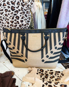 A large beige canvas tote bag with black vertical stripes and circular geometric patterns, featuring black leather handles and trim, displayed on a white wooden table.