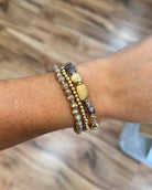 A close-up of a person's wrist wearing three stacked beaded bracelets. The bracelets include gold-toned beads, clear faceted beads, and natural stone beads in brown, gray, and yellow tones, set against a blurred wooden floor background.