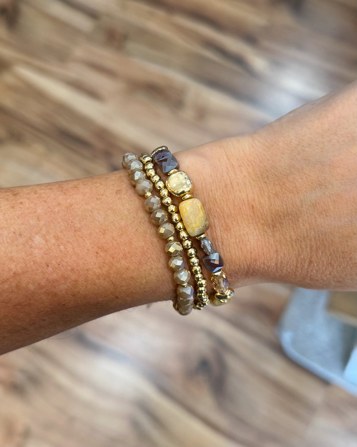A close-up of a person's wrist wearing three stacked beaded bracelets. The bracelets include gold-toned beads, clear faceted beads, and natural stone beads in brown, gray, and yellow tones, set against a blurred wooden floor background.