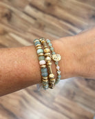 A close-up of a person's wrist wearing three beaded bracelets. The bracelets feature a mix of gold-toned spacers, light blue-green beads, pale pink beads, and a small gold elephant charm.