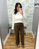 A woman takes a mirror selfie in a fitting room wearing a cream ribbed long-sleeve mock neck sweater and brown wide-leg pants with decorative gold hardware on the front pockets.