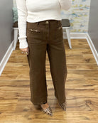A person wearing brown wide-leg corduroy pants with gold-tone horsebit pocket details, paired with a white ribbed sweater and leopard print flats, standing on a wood floor.