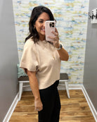A woman taking a mirror selfie wearing a beige short-sleeve knit top and black pants in a home setting with a wood floor and patterned wallpaper.