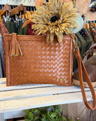 A tan woven leather clutch bag with a tassel zipper pull and wrist strap, displayed on a wooden shelf in a boutique setting with dried flowers and clothing racks in the background.