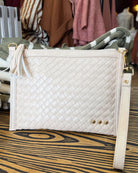 A cream-colored woven leather clutch bag with a tassel detail and a wrist strap, displayed on a wooden table with a clothing rack in the background.