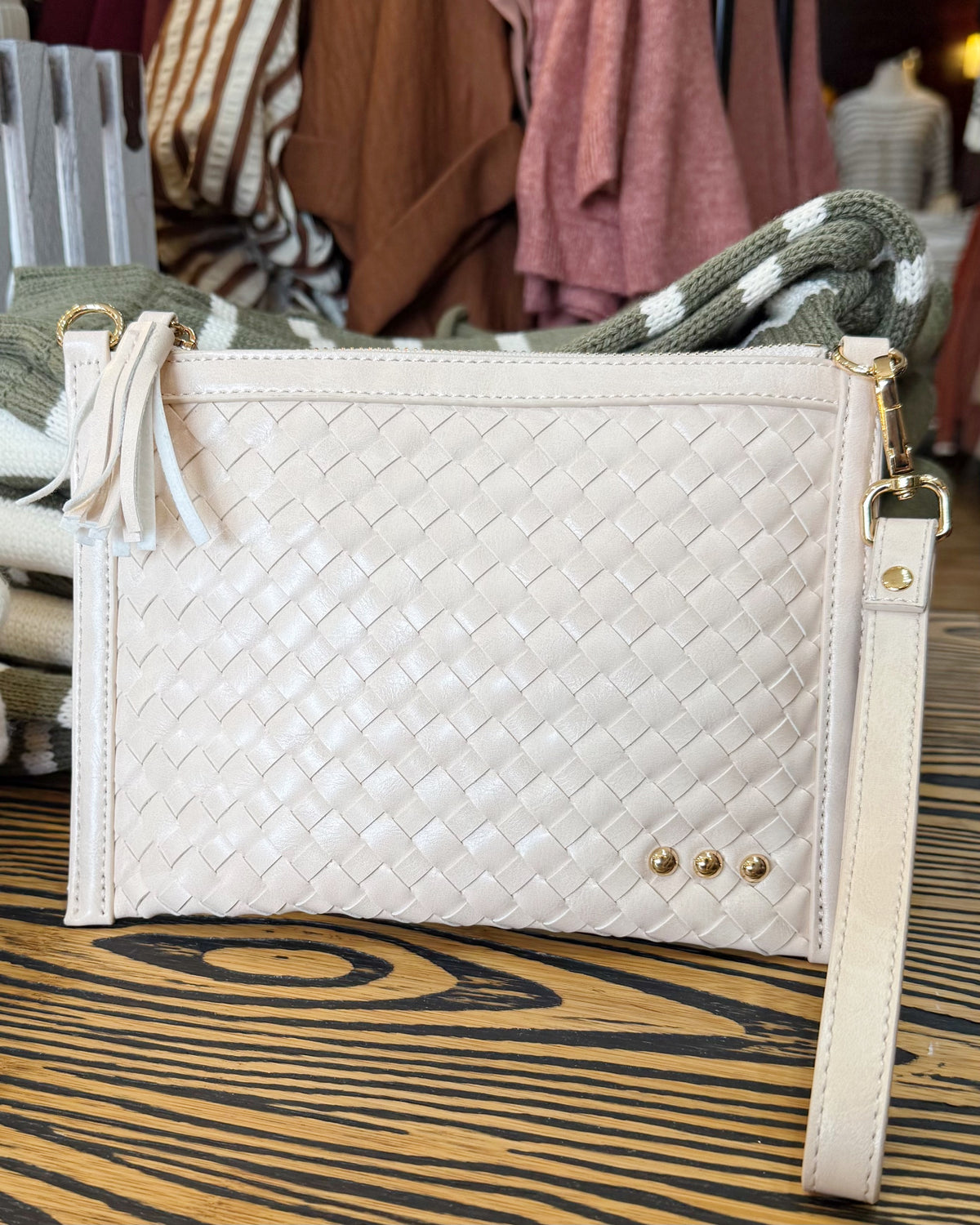 A cream-colored woven leather clutch bag with a tassel detail and a wrist strap, displayed on a wooden table with a clothing rack in the background.