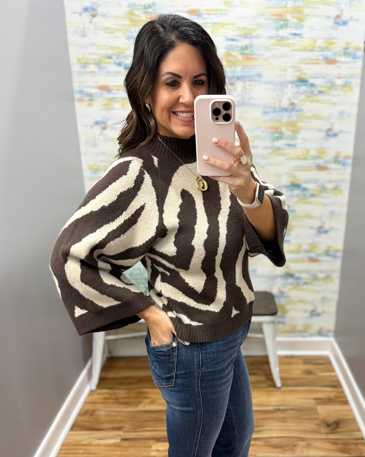 A woman takes a mirror selfie wearing a brown and cream zebra-patterned knit sweater and blue jeans in an indoor room with a wood floor and abstract wall art.