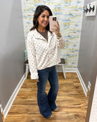 A woman takes a mirror selfie in a fitting room wearing a white zip-up jacket with black polka dots, a brown t-shirt, and blue flare jeans.