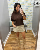 A woman takes a mirror selfie in a fitting room wearing a brown short-sleeve knit top, a beige mini skirt, and beige knee-high boots.