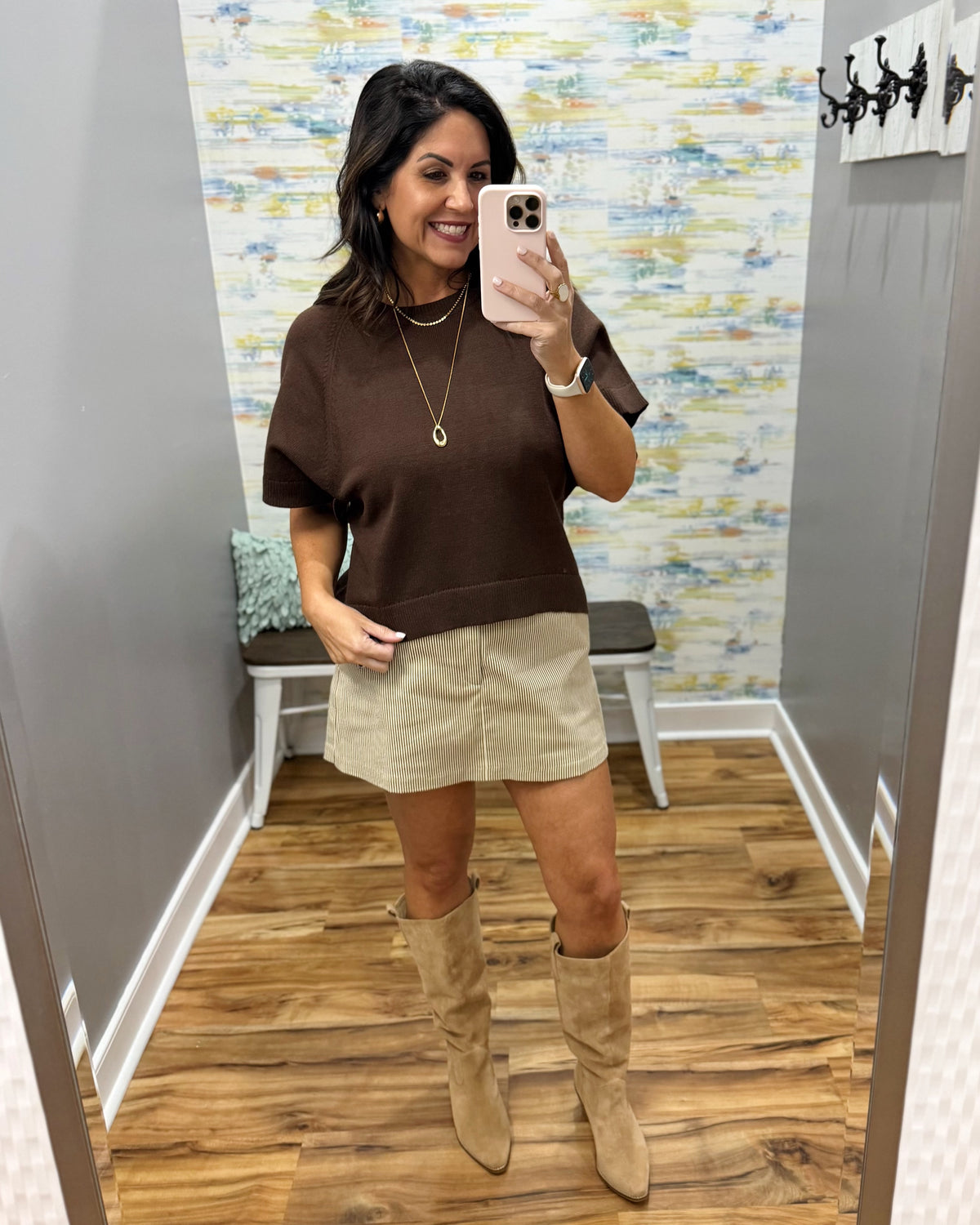 A woman takes a mirror selfie in a fitting room wearing a brown short-sleeve knit top, a beige mini skirt, and beige knee-high boots.