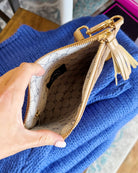 A close-up of a hand holding open a beige leather clutch or pouch with gold-tone hardware, showing a patterned interior lining and a tassel zipper pull, set against a bright blue knit fabric background.