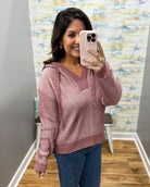 A woman takes a mirror selfie wearing a dusty rose pink ribbed knit hoodie sweater with a V-neck and drawstring hood, paired with blue jeans in an indoor setting.
