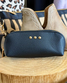 A navy blue pebbled leather zippered pouch with three gold-tone studs, displayed on a wooden slice with beige western-style ankle boots in the background.