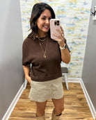 A woman takes a mirror selfie in a fitting room wearing a brown short-sleeve knit sweater, beige corduroy shorts, and beige boots.