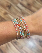 A close-up of a person's wrist wearing a stack of colorful beaded stretch bracelets with gold-toned spacer beads, set against a blurred wooden floor background.