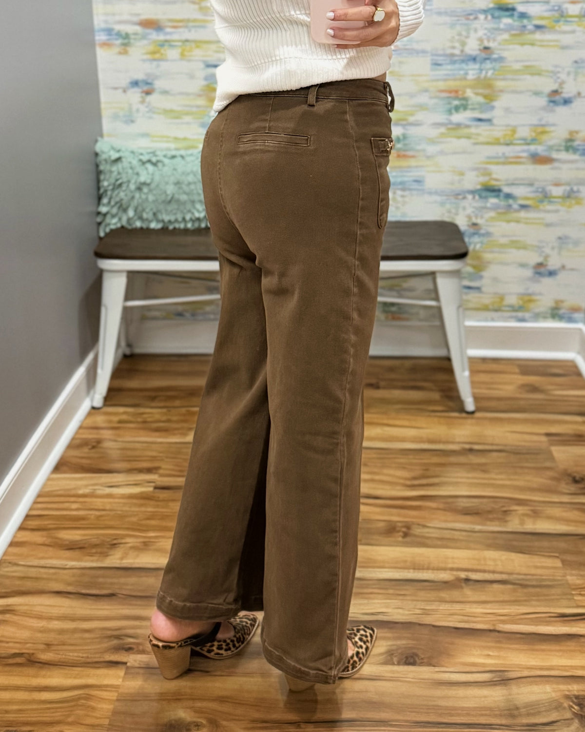 A person shown from the back wearing brown wide-leg corduroy pants, a white ribbed sweater, and leopard-print heeled mules in an indoor room with wood floors and a bench.