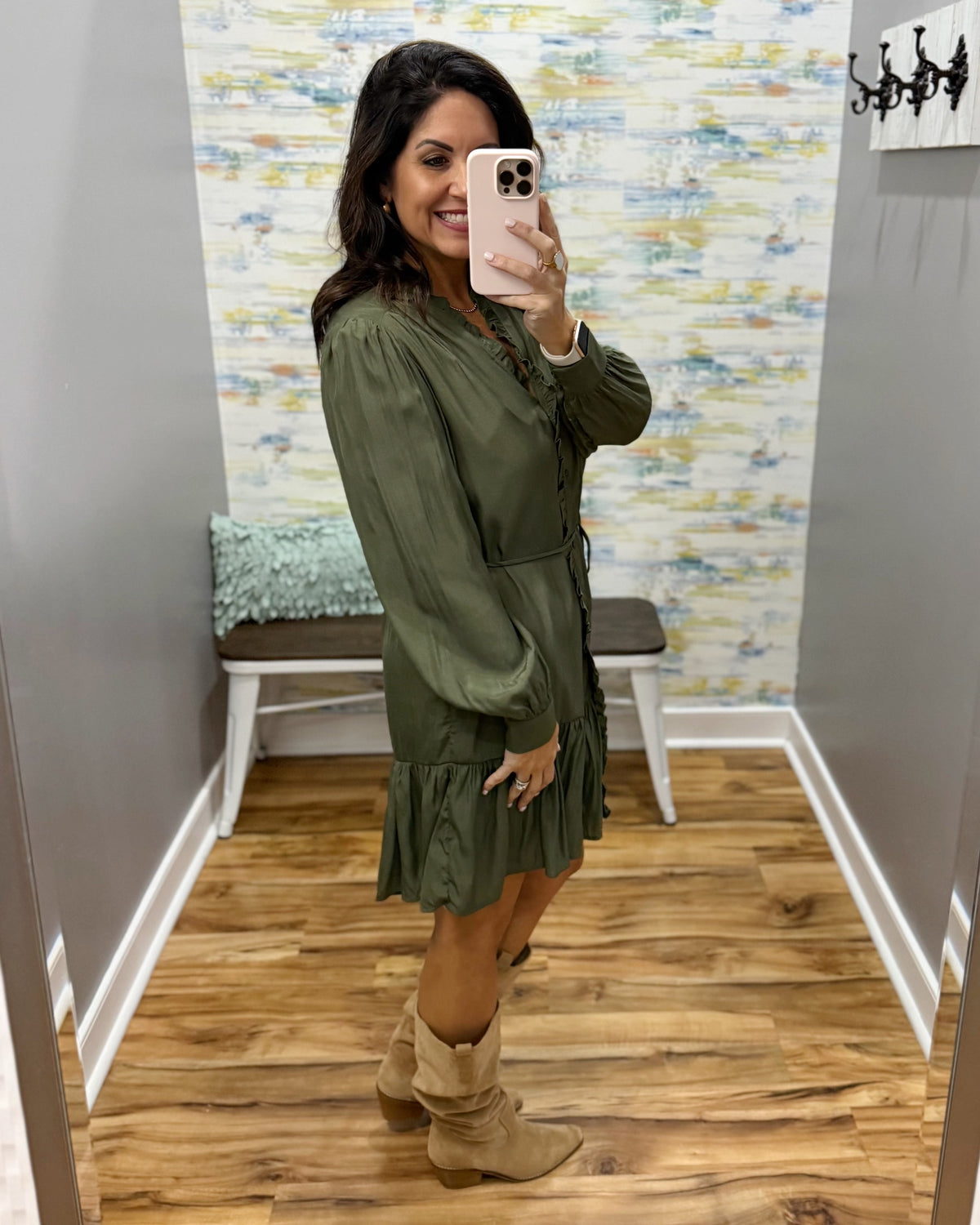 A woman takes a mirror selfie in a fitting room wearing an olive green long-sleeve mini dress with a ruffled hem and beige suede-like boots.