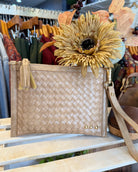 A woven beige clutch bag with a tassel zipper pull and gold-tone hardware, styled on a wooden shelf in a boutique with dried flowers and clothing racks in the background.
