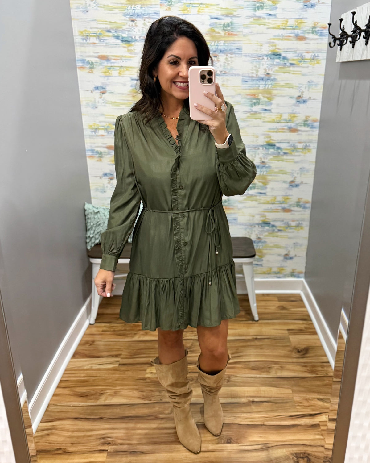 A woman takes a mirror selfie in a fitting room wearing an olive green long-sleeve mini dress with a ruffled collar and waist tie, paired with beige knee-high slouchy boots.