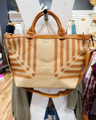 A woven tote bag with a beige and brown geometric pattern, featuring tan leather handles and a matching shoulder strap, displayed on a mannequin in a clothing store.