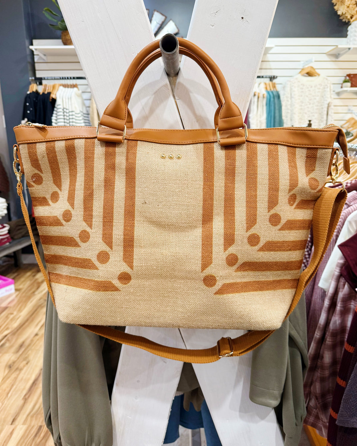 A woven tote bag with a beige and brown geometric pattern, featuring tan leather handles and a matching shoulder strap, displayed on a mannequin in a clothing store.