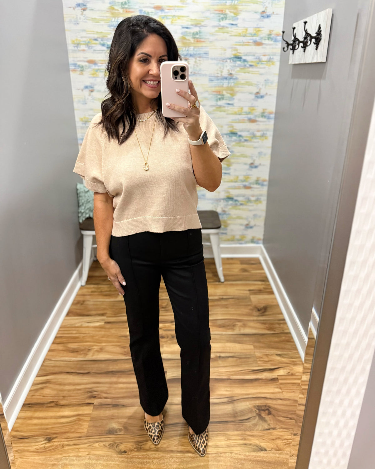 A woman takes a mirror selfie in a fitting room wearing a beige short-sleeve knit top, black pants, and leopard print heels.
