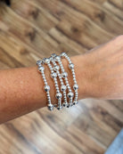 A close-up of a person's wrist wearing five stacked silver beaded bracelets, with a blurred wooden floor in the background.