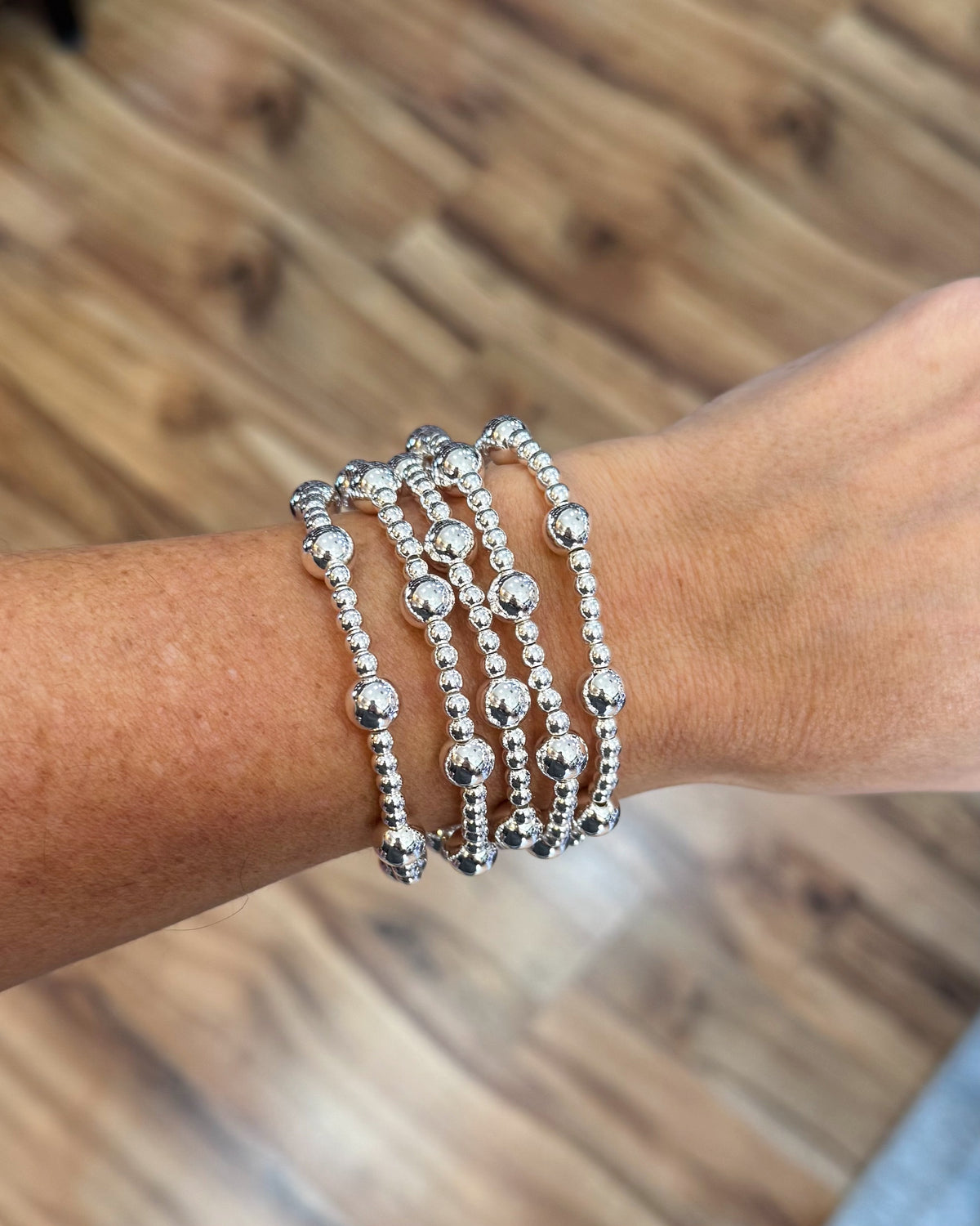 A close-up of a person's wrist wearing five stacked silver beaded bracelets, with a blurred wooden floor in the background.