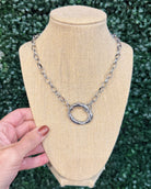 A silver-toned chain necklace with a textured circular ring pendant, displayed on a beige fabric jewelry bust. A hand with brown nail polish is holding the left side of the bust, and a green hedge wall serves as the background.